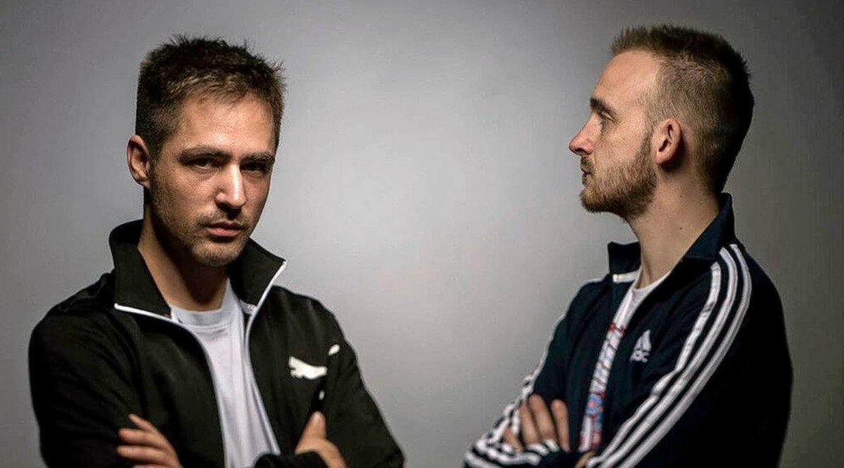 Two man on screen with the image showing them from the elbow up. Both stand with their arms crossed wearing black with white striped track suits. Both stand arms crossed, one looking straight ahead, the other stares to the left and looking at the other man.